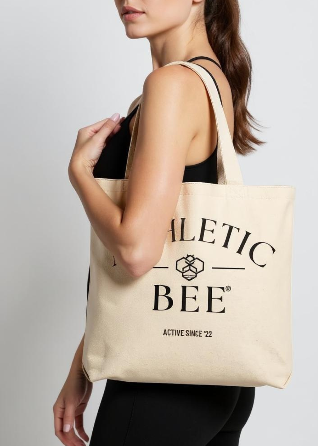 Person carrying a beige tote bag with 'Alettic Bee' branding on a light gray background